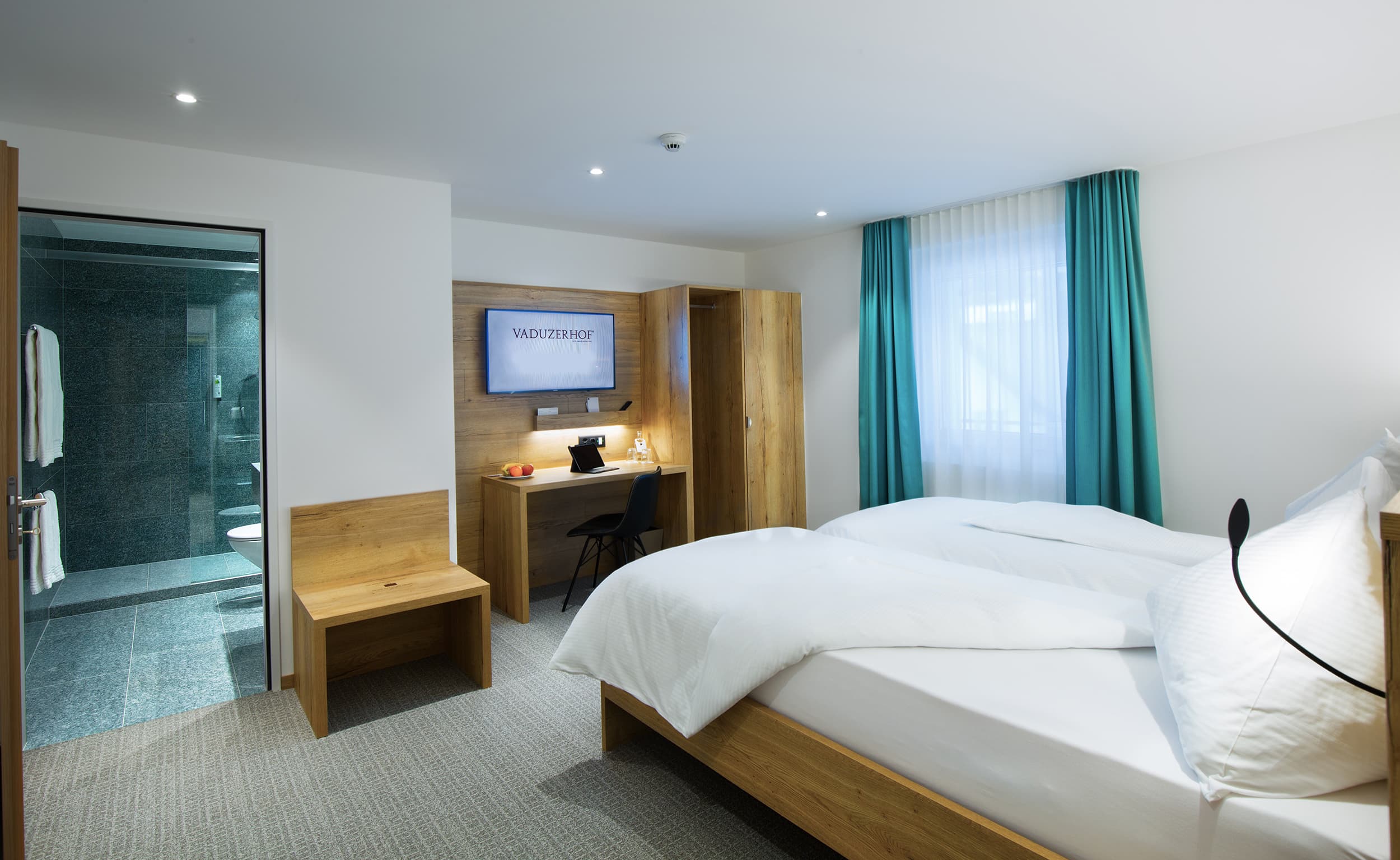 A modern double and twin room in the Vaduzerhof with wood interior, desk, TV, and adjoining bathroom.