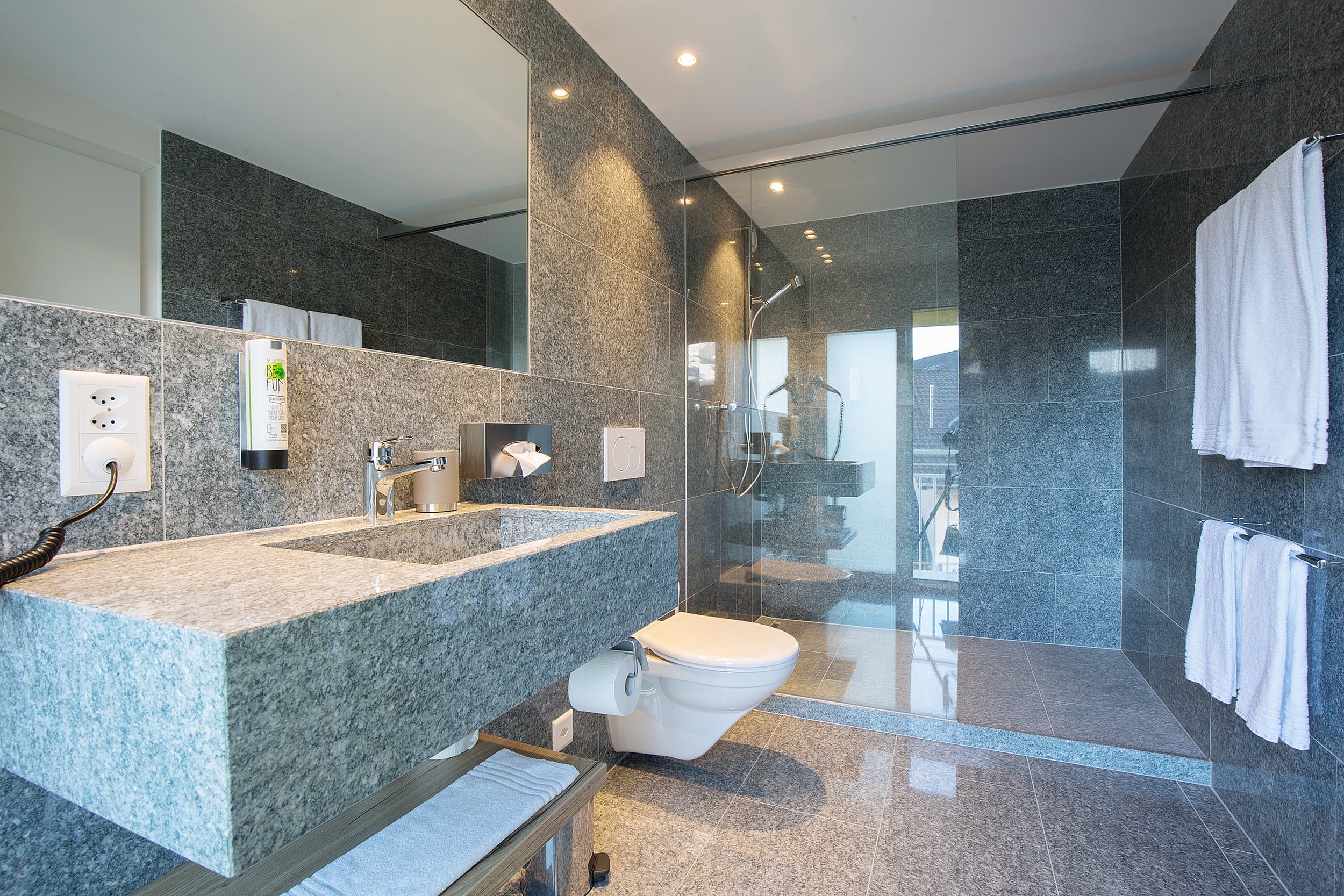 A modern bathroom in the Junior Suite of the Vaduzerhof by B Smart with gray tiles, a sink, a toilet, and a glass shower.