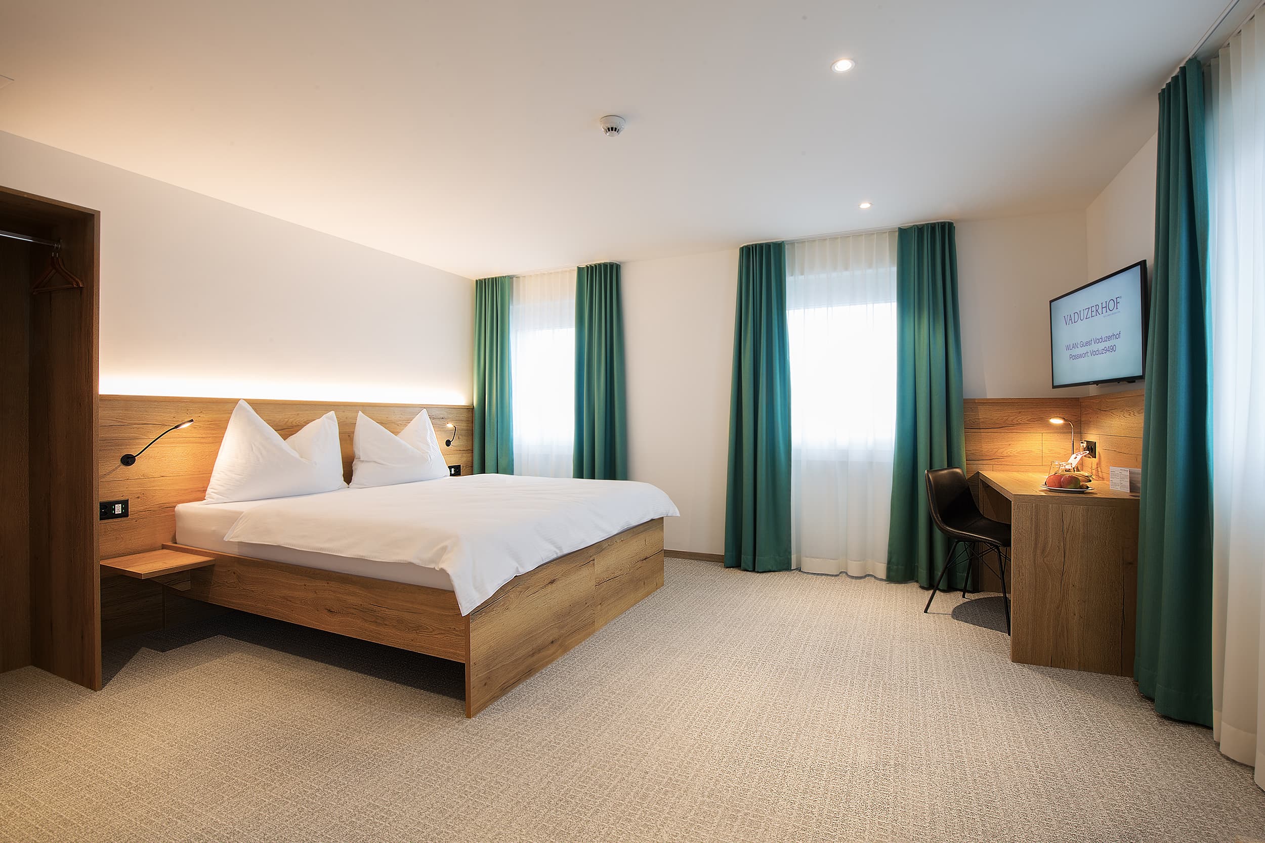 A comfortably furnished double room in the Vaduzerhof with a large bed, desk, chair, carpeted floor, and windows with turquoise curtains.