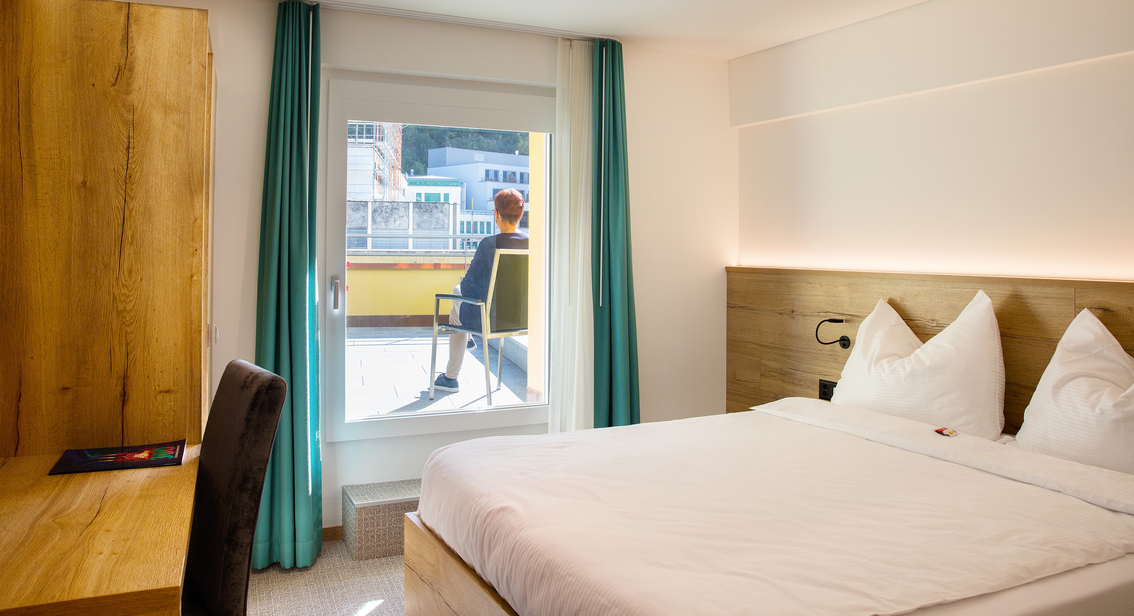 A double room in the Vaduzerhof with a large bed a wooden desk and a person sitting on a balcony
